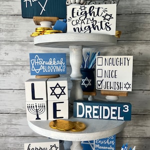 May include: A tiered white tray decorated for Hanukkah with blue and white signs, gold coins, and a menorah. The signs say "Happy Hanukkah", "Eight Crazy Nights", "Hanukkah Blessings", "Naughty Nice Jewish", "Dreidel 3", and "Happy Challah Days".