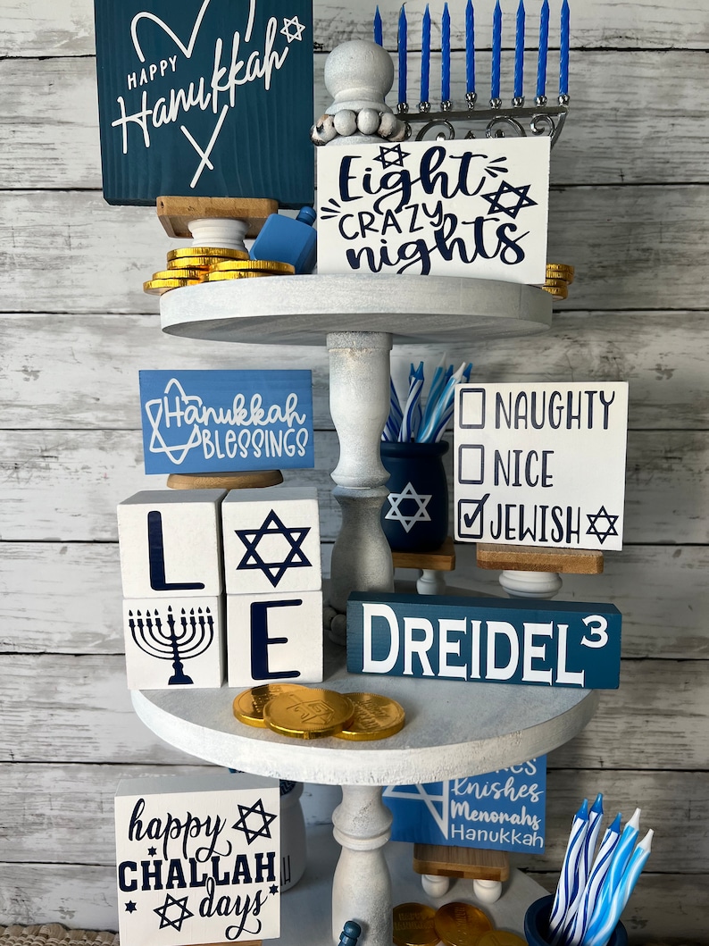 May include: A tiered white tray decorated for Hanukkah with blue and white signs, gold coins, and a menorah. The signs say "Happy Hanukkah", "Eight Crazy Nights", "Hanukkah Blessings", "Naughty Nice Jewish", "Dreidel 3", and "Happy Challah Days".