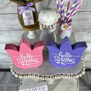 K&ouml;nnte beinhalten: Zwei Holzschildchen mit dem Text "hello spring" in wei&szlig;en Buchstaben. Die Schilder sind in rosa und lila bemalt und in Form von Tulpen. Die Schilder stehen auf einem wei&szlig;en, mehrst&ouml;ckigen St&auml;nder mit einem Schild, auf dem "flower market" steht, und einer kleinen Vase mit Blumen und einem Schild, auf dem "fresh flowers 25c" steht.