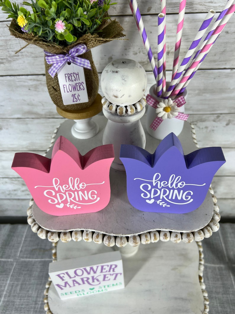 K&ouml;nnte beinhalten: Zwei Holzschildchen mit dem Text "hello spring" in wei&szlig;en Buchstaben. Die Schilder sind in rosa und lila bemalt und in Form von Tulpen. Die Schilder stehen auf einem wei&szlig;en, mehrst&ouml;ckigen St&auml;nder mit einem Schild, auf dem "flower market" steht, und einer kleinen Vase mit Blumen und einem Schild, auf dem "fresh flowers 25c" steht.