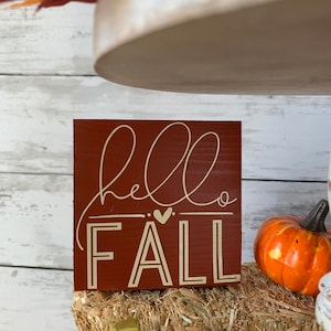May include: A small wooden sign with the words "hello fall" written in white script on a red background. The sign is sitting on a bale of hay with a small orange pumpkin in the background.