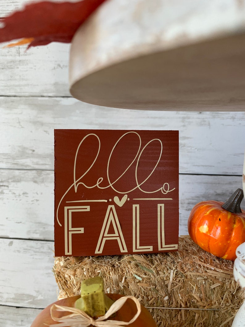 May include: A small wooden sign with the words "hello fall" written in white script on a red background. The sign is sitting on a bale of hay with a small orange pumpkin in the background.