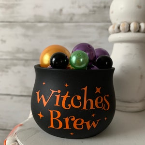 May include: Black cauldron with the text "Witches Brew" in orange lettering. The cauldron is filled with purple, green, gold, and black beads.
