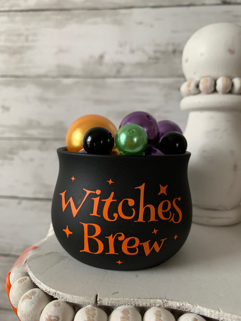 May include: Black cauldron with the text "Witches Brew" in orange lettering. The cauldron is filled with purple, green, gold, and black beads.