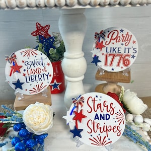 May include: Three round, white, wooden signs with red, white, and blue decorations and text. The signs say "Sweet Land of Liberty", "Party Like It's 1776", and "Stars and Stripes".