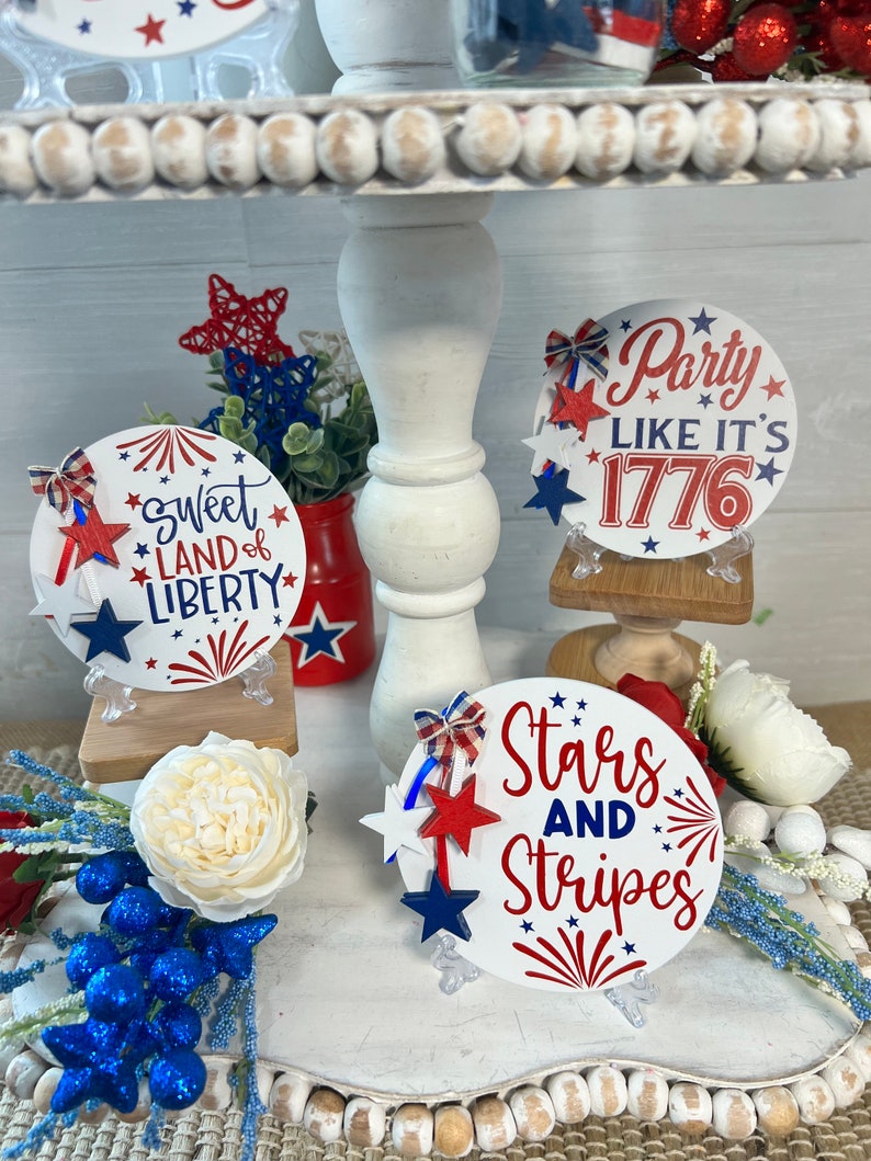 May include: Three round, white, wooden signs with red, white, and blue decorations and text. The signs say "Sweet Land of Liberty", "Party Like It's 1776", and "Stars and Stripes".