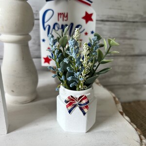 Pode incluir: Um pote de cer&acirc;mica branco com um la&ccedil;o vermelho, branco e azul est&aacute; sobre uma superf&iacute;cie branca. O pote est&aacute; cheio de um pequeno buqu&ecirc; de flores artificiais azuis e verdes. Um letreiro de madeira com o texto "my home" &eacute; vis&iacute;vel no fundo.
