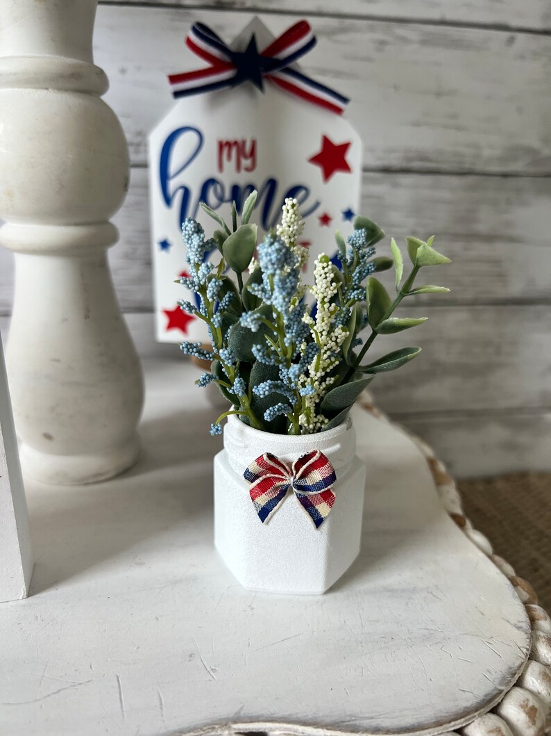 Pode incluir: Um pote de cer&acirc;mica branco com um la&ccedil;o vermelho, branco e azul est&aacute; sobre uma superf&iacute;cie branca. O pote est&aacute; cheio de um pequeno buqu&ecirc; de flores artificiais azuis e verdes. Um letreiro de madeira com o texto "my home" &eacute; vis&iacute;vel no fundo.