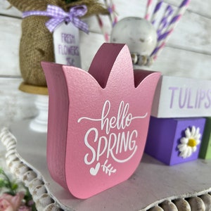 K&ouml;nnte beinhalten: Ein rosafarbenes, tulpenf&ouml;rmiges Schild aus Holz mit dem Text "hello spring" in wei&szlig;en Buchstaben. Das Schild steht auf einem wei&szlig;en, mehrst&ouml;ckigen Tablett mit einem wei&szlig;en Perlenrand.