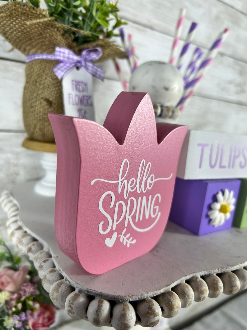 K&ouml;nnte beinhalten: Ein rosafarbenes, tulpenf&ouml;rmiges Schild aus Holz mit dem Text "hello spring" in wei&szlig;en Buchstaben. Das Schild steht auf einem wei&szlig;en, mehrst&ouml;ckigen Tablett mit einem wei&szlig;en Perlenrand.