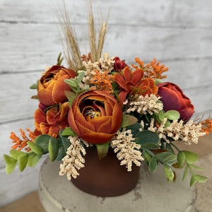 May include: A fall-themed floral arrangement in a brown vase. The arrangement features large, orange-red peony-like flowers, smaller orange blooms, and green foliage. Tall wheat stalks add height and texture to the display.