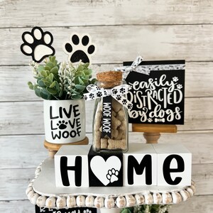 May include: A tiered tray decorated with dog-themed home decor. The tray features a white ceramic mug with the words "Live Love Woof", a glass jar filled with dog treats, and three white wooden blocks spelling "HOME" with a paw print heart in the center. The tray also includes two black and white signs with the text "Easily Distracted by Dogs" and "Is My Favorite Breed".