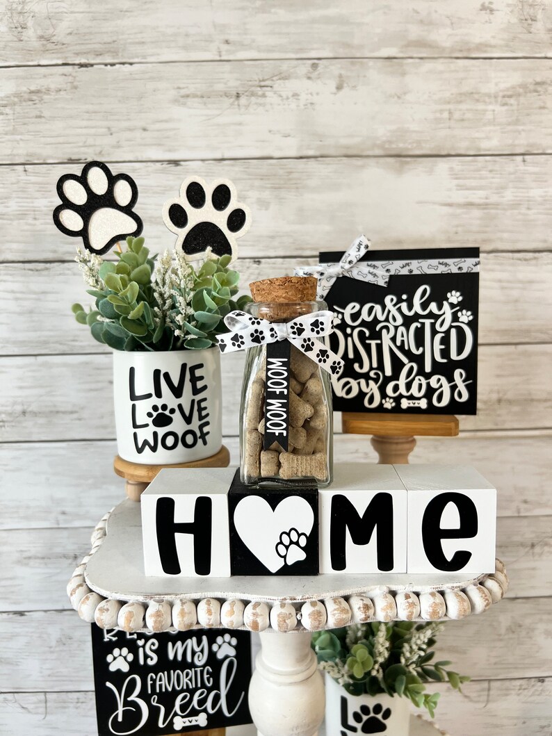 May include: A tiered tray decorated with dog-themed home decor. The tray features a white ceramic mug with the words "Live Love Woof", a glass jar filled with dog treats, and three white wooden blocks spelling "HOME" with a paw print heart in the center. The tray also includes two black and white signs with the text "Easily Distracted by Dogs" and "Is My Favorite Breed".