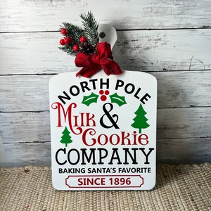 May include: White wooden sign with a red bow and green pine sprigs. The sign reads "North Pole Milk & Cookie Company Baking Santa's Favourite Since 1896".