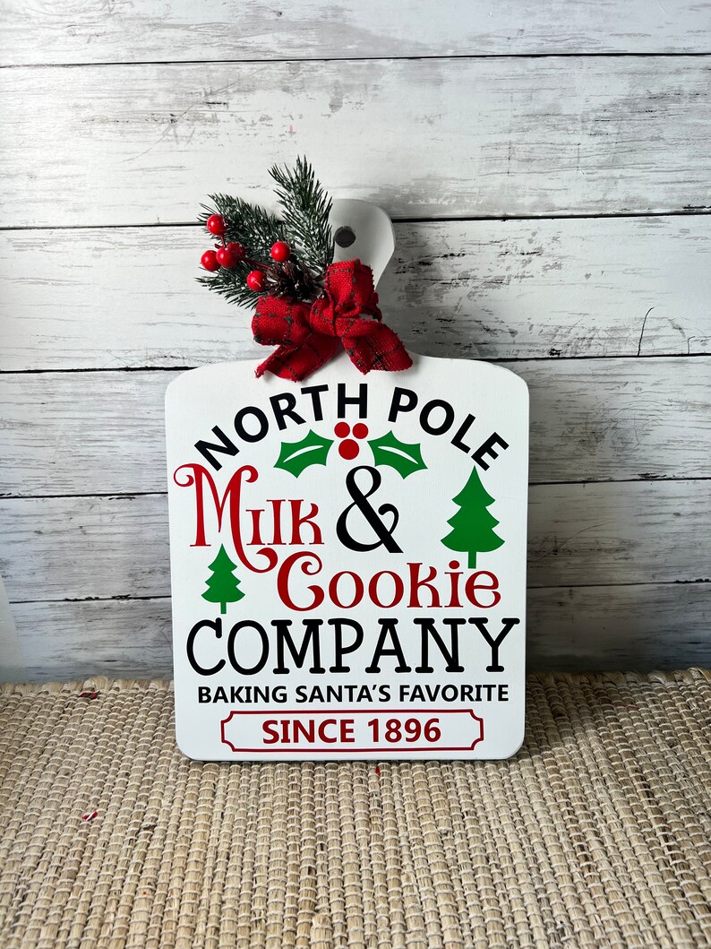 May include: White wooden sign with a red bow and green pine sprigs. The sign reads "North Pole Milk & Cookie Company Baking Santa's Favourite Since 1896".