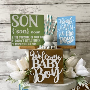K&ouml;nnte beinhalten: Ein dreist&ouml;ckiger Kuchenst&auml;nder mit drei Holzschilden. Das oberste Schild ist gr&uuml;n und sagt "SON {son} noun 1. THE SUNSHINE OF YOUR DAY 2. DADDY'S LITTLE HELPER 3. MAMA'S LITTLE MAN". Das mittlere Schild ist braun und sagt "Welcome BABY Boy". Das unterste Schild ist braun und sagt "Mommy's Little Man Daddy's".