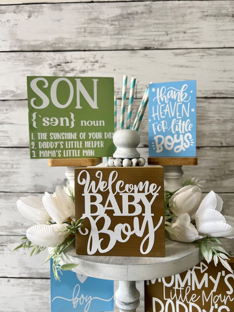 K&ouml;nnte beinhalten: Ein dreist&ouml;ckiger Kuchenst&auml;nder mit drei Holzschilden. Das oberste Schild ist gr&uuml;n und sagt "SON {son} noun 1. THE SUNSHINE OF YOUR DAY 2. DADDY'S LITTLE HELPER 3. MAMA'S LITTLE MAN". Das mittlere Schild ist braun und sagt "Welcome BABY Boy". Das unterste Schild ist braun und sagt "Mommy's Little Man Daddy's".