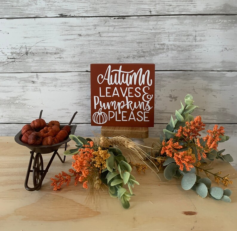 May include: A small wooden sign with a red background and white lettering that reads "Autumn Leaves & Pumpkins Please". The sign is sitting on a wooden stand and is surrounded by small orange and green plants.