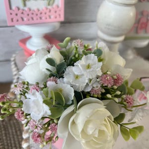 May include: A small pink planter with a bouquet of white and pink flowers. The flowers include white roses, small white flowers, and pink buds.