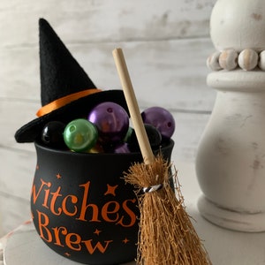 May include: A black cauldron with the text "Witches Brew" in orange letters, filled with colorful beads and topped with a black witch's hat. A miniature broom with a brown handle and a black and white striped band is next to the cauldron.