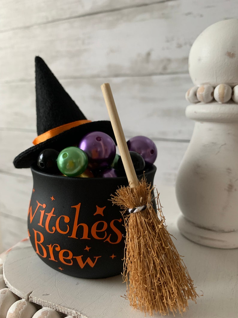 May include: A black cauldron with the text "Witches Brew" in orange letters, filled with colorful beads and topped with a black witch's hat. A miniature broom with a brown handle and a black and white striped band is next to the cauldron.