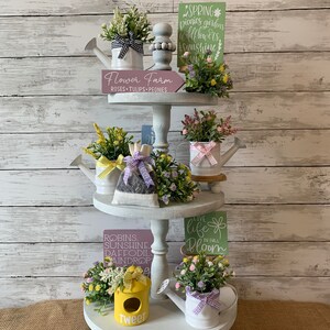 May include: A three-tiered, white, decorative tray with spring-themed decor. The tray holds miniature watering cans with faux flowers, signs with floral and seasonal text, and a yellow birdhouse. The text includes "Flower Farm" and "Tweet".