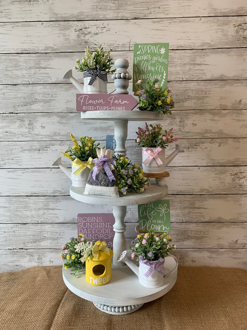 May include: A three-tiered, white, decorative tray with spring-themed decor. The tray holds miniature watering cans with faux flowers, signs with floral and seasonal text, and a yellow birdhouse. The text includes "Flower Farm" and "Tweet".