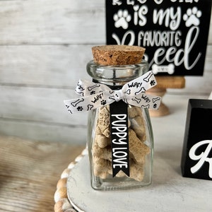 May include: A glass jar filled with dog biscuits, tied with a white ribbon with paw prints and the word "Woof!" printed on it. A black banner with the text "Puppy Love" is attached to the ribbon. The jar is sitting on a white tiered stand with a black sign that reads "Rescued is my favorite breed".