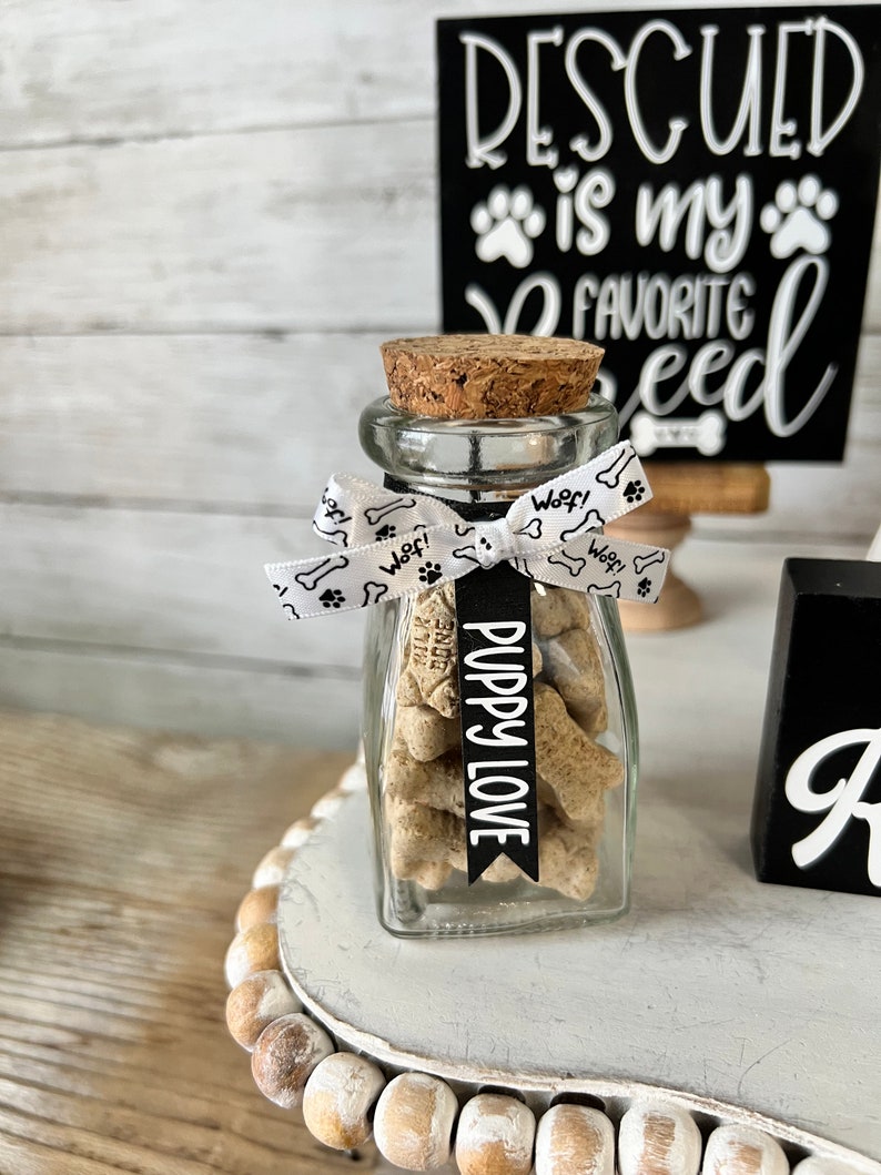May include: A glass jar filled with dog biscuits, tied with a white ribbon with paw prints and the word "Woof!" printed on it. A black banner with the text "Puppy Love" is attached to the ribbon. The jar is sitting on a white tiered stand with a black sign that reads "Rescued is my favorite breed".