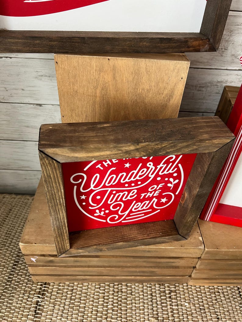 May include: A red wooden sign with white lettering that reads "The Most Wonderful Time of the Year". The sign is framed in dark wood and is sitting on a wooden shelf.