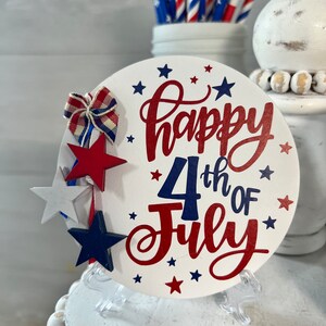 May include: A white round wooden sign with the text "Happy 4th of July" in red and blue lettering. The sign is decorated with red, white, and blue stars and a red, white, and blue bow. The sign is on a clear plastic stand.