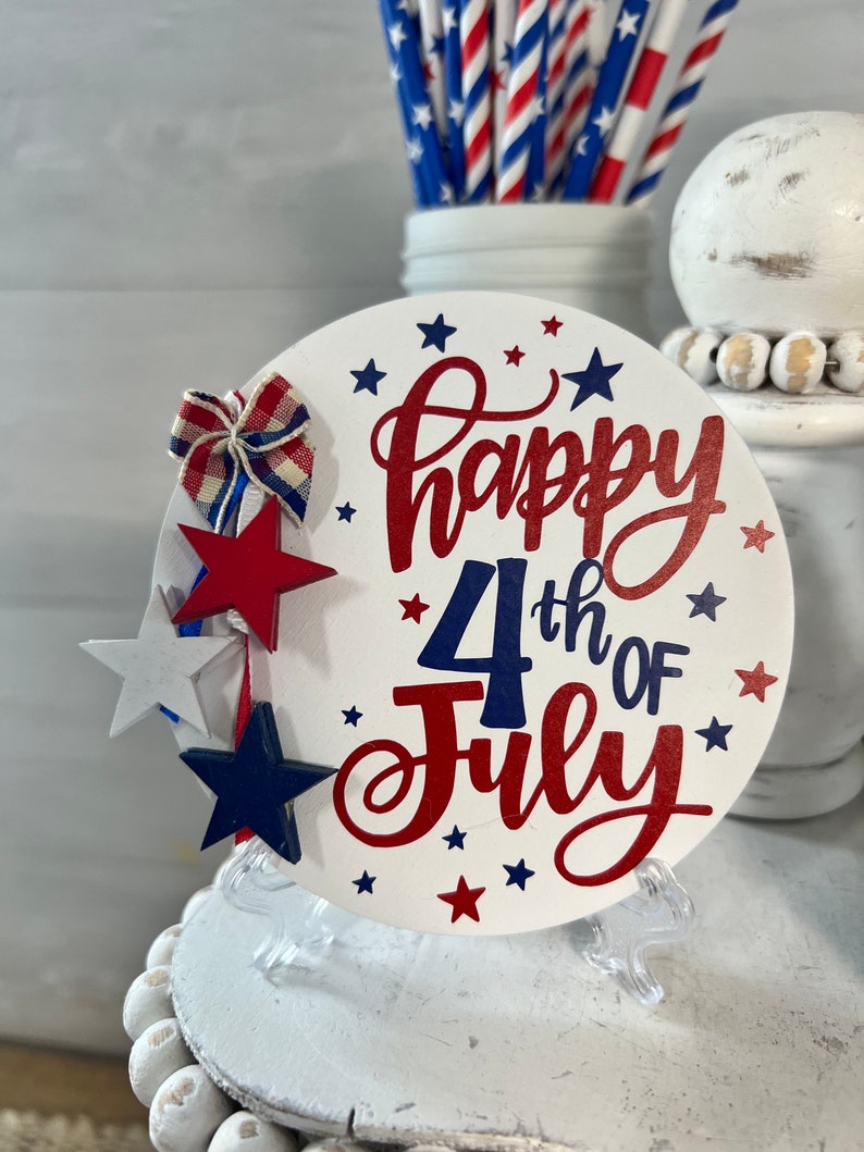 May include: A white round wooden sign with the text "Happy 4th of July" in red and blue lettering. The sign is decorated with red, white, and blue stars and a red, white, and blue bow. The sign is on a clear plastic stand.