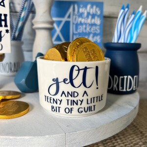 May include: A white ceramic mug with blue lettering that reads "gelt! and a teeny tiny little bit of guilt". The mug is filled with gold chocolate coins.