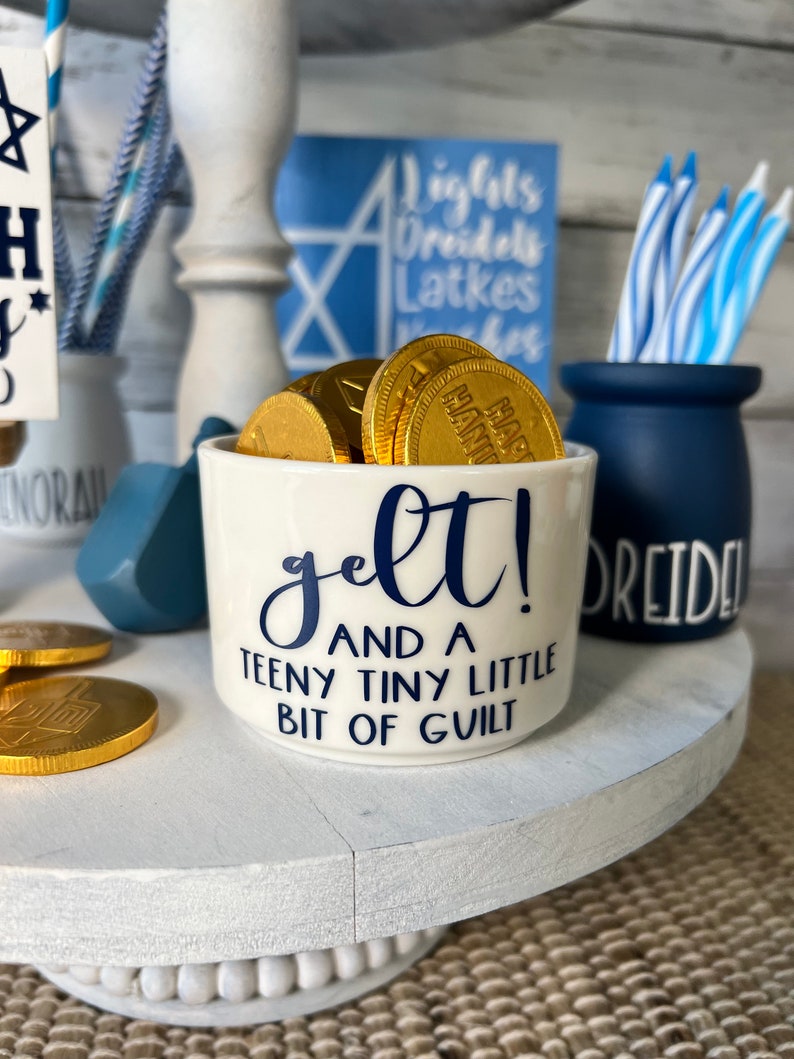 May include: A white ceramic mug with blue lettering that reads "gelt! and a teeny tiny little bit of guilt". The mug is filled with gold chocolate coins.