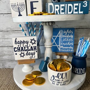 May include: A white tiered tray decorated for Hanukkah with blue and white wooden blocks spelling "DREIDEL", a blue and white dreidel, a white mug with gold coins and the text "gelt! and a teeny tiny bit of guilt", a blue and white striped straw holder, and a sign that says "happy CHALLAH days".