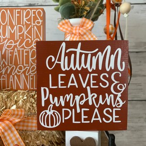 May include: A brown wooden sign with white lettering that reads "Autumn Leaves & Pumpkins Please". The sign is decorated with a white pumpkin graphic. The sign is sitting on a shelf with other fall-themed decorations.