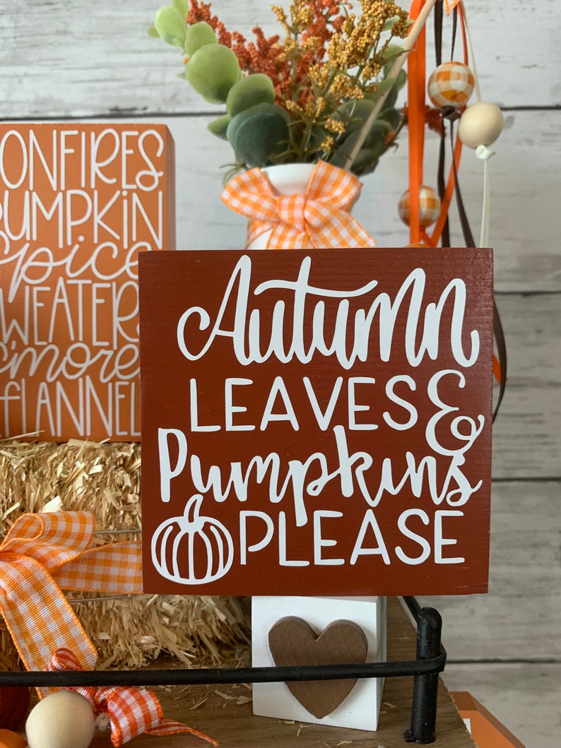 May include: A brown wooden sign with white lettering that reads "Autumn Leaves & Pumpkins Please". The sign is decorated with a white pumpkin graphic. The sign is sitting on a shelf with other fall-themed decorations.