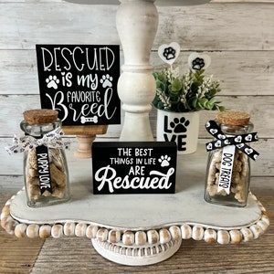May include: A white tiered tray with a black sign that reads "Rescued is my favorite breed". There are two glass jars filled with dog treats, one labeled "Puppy Love" and the other "Dog Treats". The tray is decorated with white beads and a small potted plant.