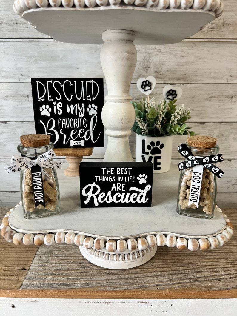 May include: A white tiered tray with a black sign that reads "Rescued is my favorite breed". There are two glass jars filled with dog treats, one labeled "Puppy Love" and the other "Dog Treats". The tray is decorated with white beads and a small potted plant.