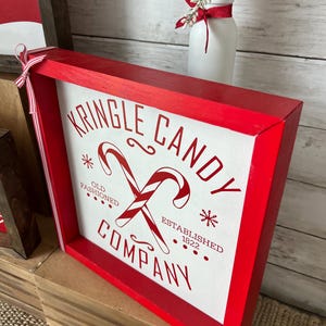 May include: Red wooden framed sign with white background and red text that reads "Kringle Candy Company" with a red and white candy cane graphic and the text "Old Fashioned" and "Established 1822".