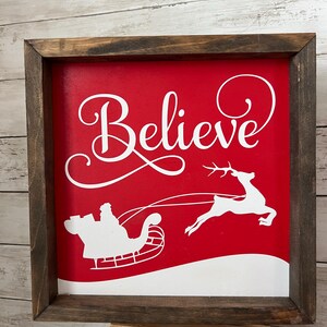 May include: A red wooden sign with a white frame. The sign says "Believe" in a white cursive font. There is a white silhouette of Santa Claus in his sleigh pulled by reindeer on the sign.