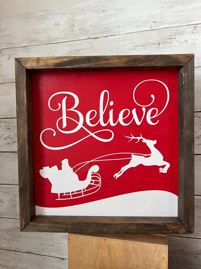 May include: A red wooden sign with a white frame. The sign says "Believe" in a white cursive font. There is a white silhouette of Santa Claus in his sleigh pulled by reindeer on the sign.