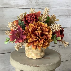 May include: A fall-themed floral arrangement featuring a large, orange dahlia as the focal point. The arrangement includes burgundy, orange, and cream-colored flowers and greenery, all nestled in a small, woven basket. The flowers sit on a small, round, gray stool.