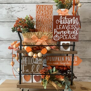 May include: A black metal cart with two shelves decorated for fall. The top shelf has a hay bale, a white vase with orange and white checkered ribbon, and three wooden signs with the text "Bonfires, Pumpkin Spice, Sweaters, Smores, Flannels", "Autumn Leaves & Pumpkins Please", and "Hello Fall". The bottom shelf has a wooden sign with the text "Pumpkin Patch, corn maze, hayrides, cider", a garland of orange and white checkered beads, and a small bouquet of fall foliage.