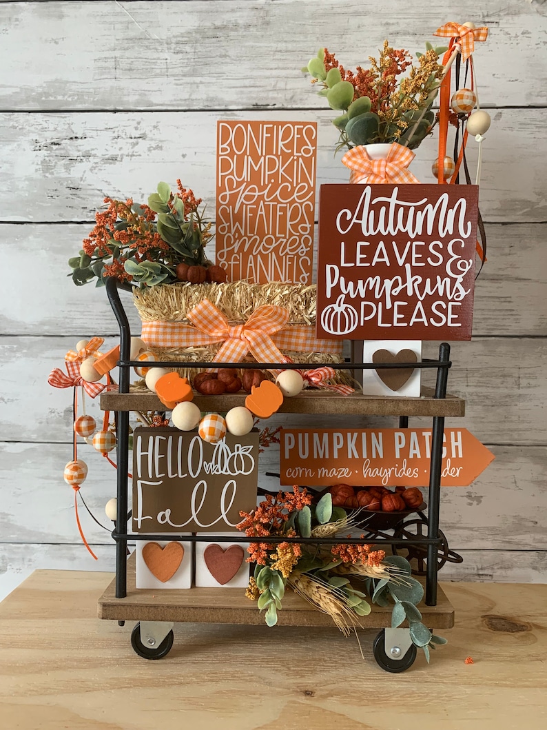 May include: A black metal cart with two shelves decorated for fall. The top shelf has a hay bale, a white vase with orange and white checkered ribbon, and three wooden signs with the text "Bonfires, Pumpkin Spice, Sweaters, Smores, Flannels", "Autumn Leaves & Pumpkins Please", and "Hello Fall". The bottom shelf has a wooden sign with the text "Pumpkin Patch, corn maze, hayrides, cider", a garland of orange and white checkered beads, and a small bouquet of fall foliage.