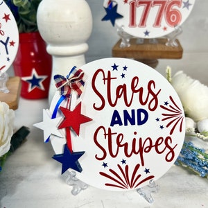 May include: White round wooden sign with the text "Stars and Stripes" in red and blue lettering. The sign has red and blue stars and a red firework design. The sign is on a white stand.