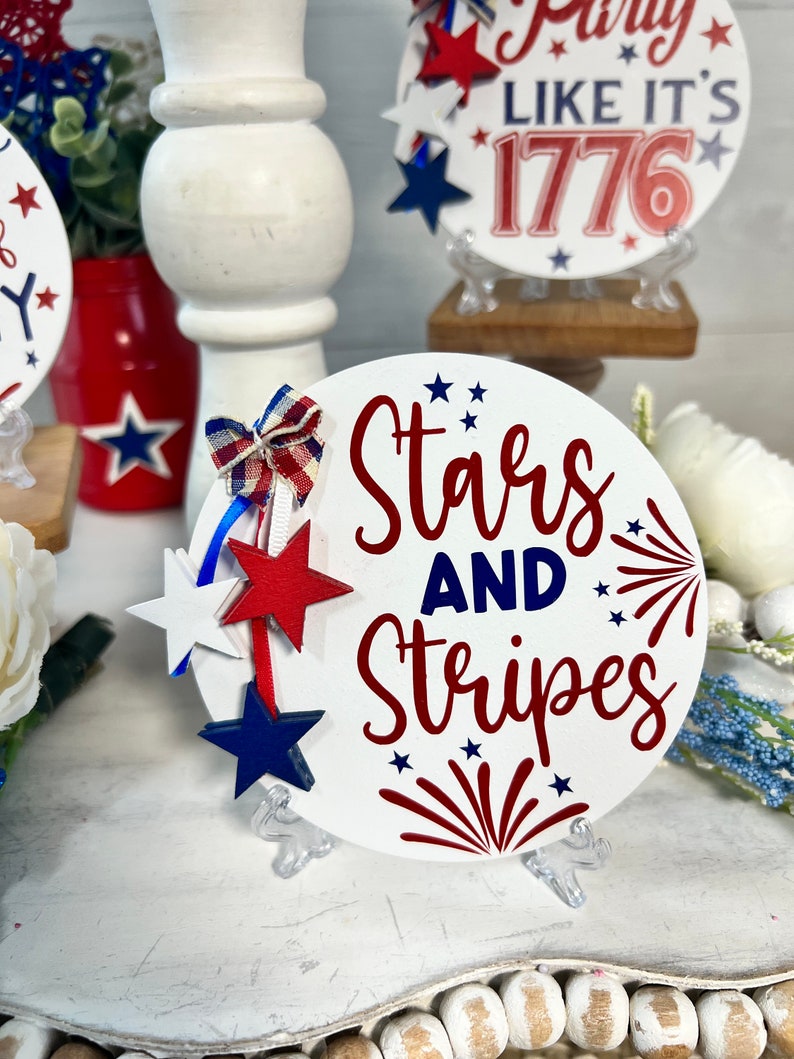 May include: White round wooden sign with the text "Stars and Stripes" in red and blue lettering. The sign has red and blue stars and a red firework design. The sign is on a white stand.