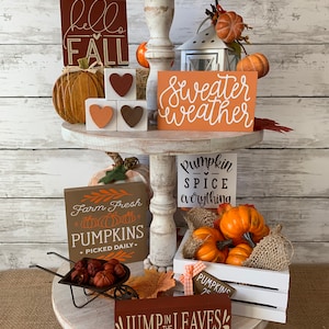 May include: A tiered tray decorated with fall-themed signs and pumpkins. The signs read "hello fall", "sweater weather", "pumpkin spice everything", "farm fresh pumpkins", and "jump in the leaves".