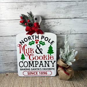 May include: White wooden sign with the text "North Pole Milk & Cookie Company Baking Santa's Favourite Since 1896". The sign has a red bow and green holly with red berries on top. A small potted artificial Christmas tree is to the right of the sign.