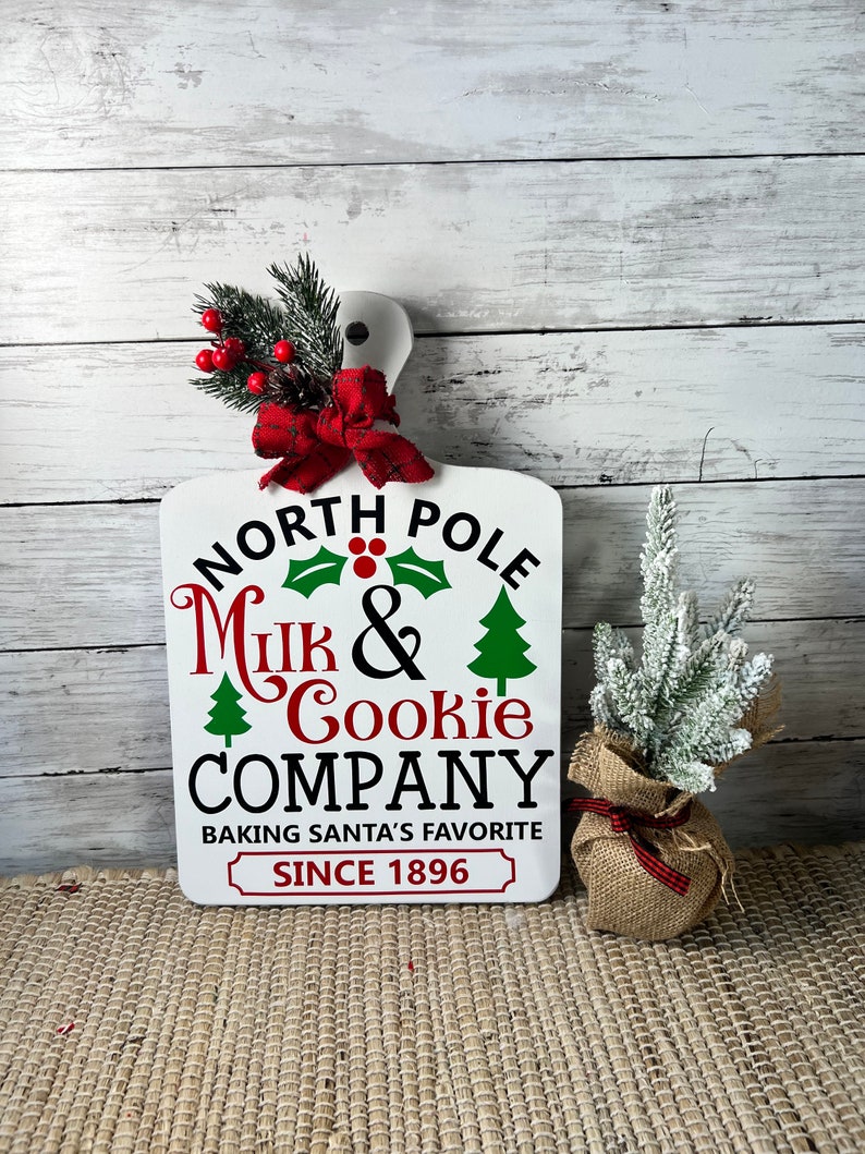 May include: White wooden sign with the text "North Pole Milk & Cookie Company Baking Santa's Favourite Since 1896". The sign has a red bow and green holly with red berries on top. A small potted artificial Christmas tree is to the right of the sign.